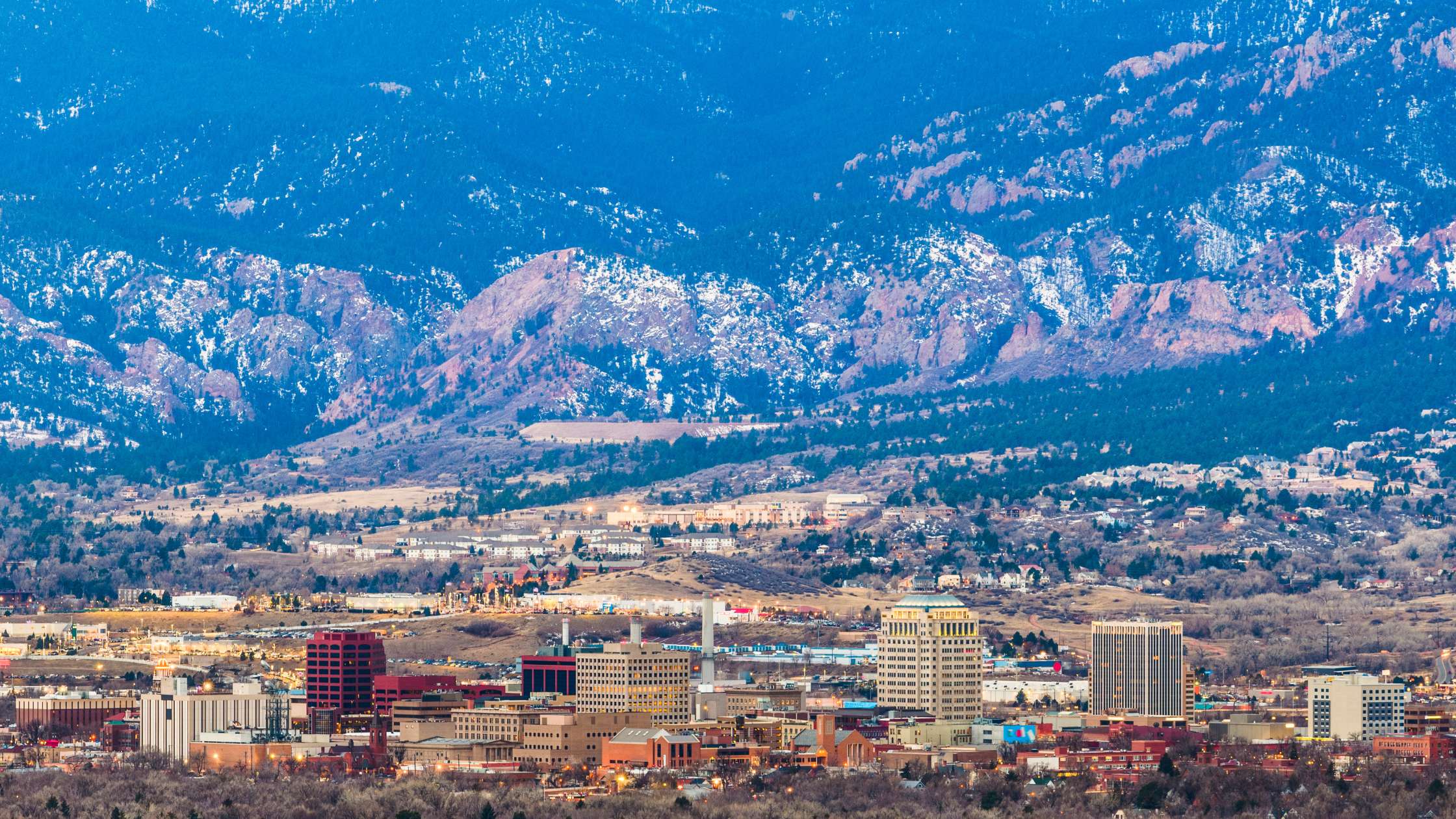 The Colorado Springs skyline at dusk, set against the snow-dusted foothills of the Rocky Mountains
